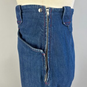 May include: High-waisted dark blue denim jeans featuring a prominent silver-toned metal zipper closure running vertically along the side. The garment includes belt loops, a single silver button at the waist, and visible red stitching accents.