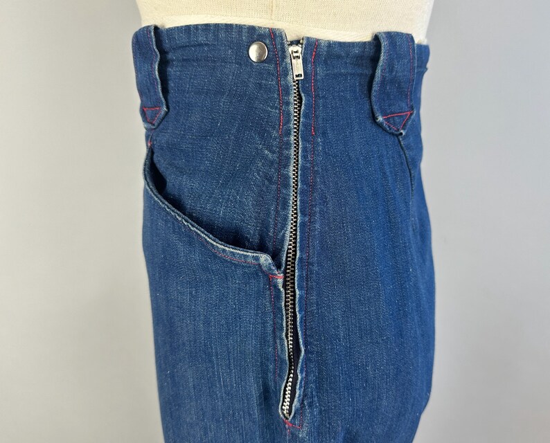 May include: High-waisted dark blue denim jeans featuring a prominent silver-toned metal zipper closure running vertically along the side. The garment includes belt loops, a single silver button at the waist, and visible red stitching accents.