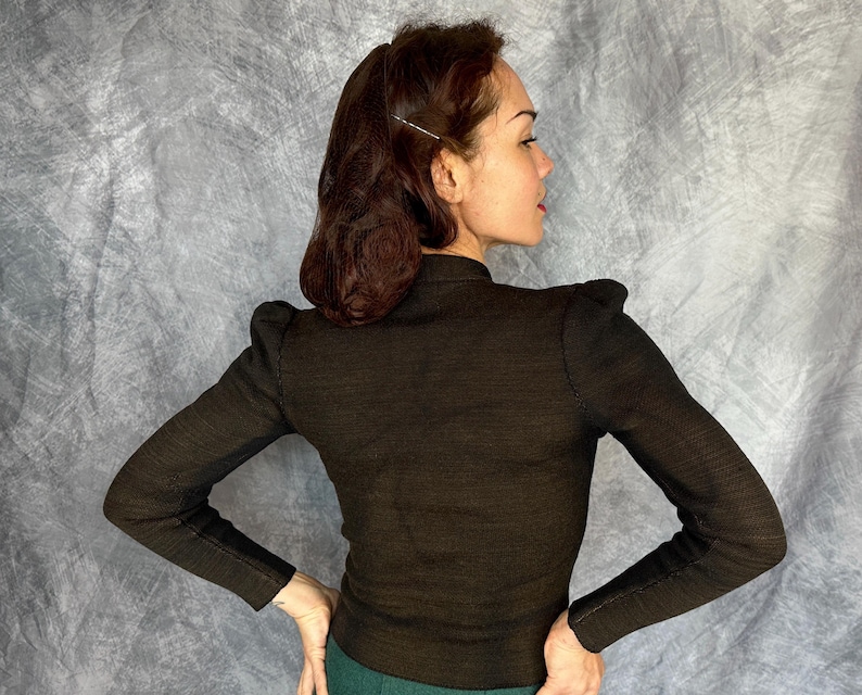 May include: A dark brown long-sleeved sweater with a high neckline and shoulder pads. The sweater has a textured appearance and is fitted. The person is wearing green pants. The background is a mottled gray.