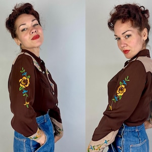 May include: A long-sleeve button-down shirt in dark brown with tan shoulder yokes and cuffs. The sleeves feature colourful floral embroidery in yellow, green, and blue. The shirt is paired with blue denim jeans featuring a button-front closure.
