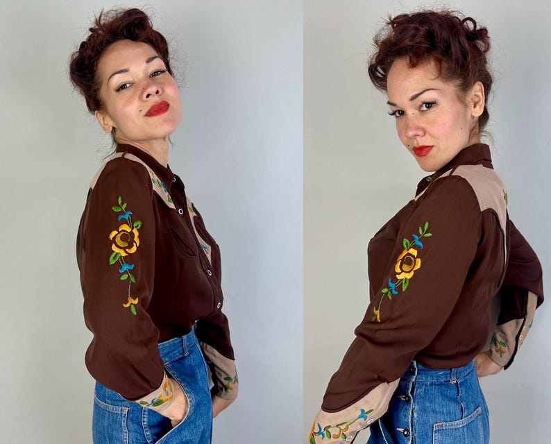 May include: A long-sleeve button-down shirt in dark brown with tan shoulder yokes and cuffs. The sleeves feature colourful floral embroidery in yellow, green, and blue. The shirt is paired with blue denim jeans featuring a button-front closure.
