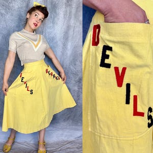 May include: A bright yellow A-line skirt featuring two large patch pockets. The left pocket has the word DEVILS appliqued in red and black felt letters, while the right pocket features the word BERRIES in red and black felt lettering.