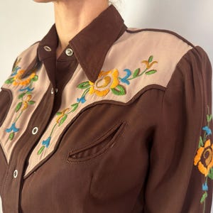 May include: A brown long-sleeve shirt featuring tan panels on the chest and shoulders. The shirt includes silver snap buttons and vibrant floral embroidery in yellow, green, and blue on the front panels and along the sleeve.