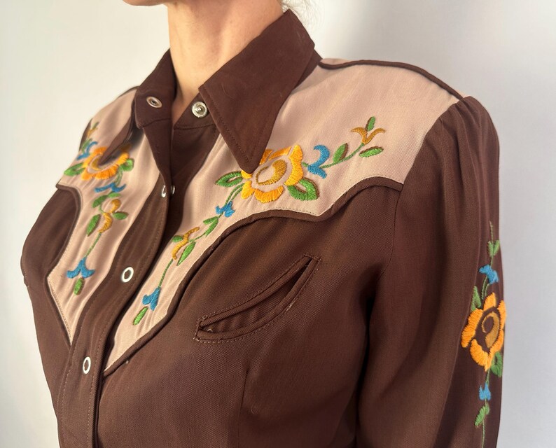 May include: A brown long-sleeve shirt featuring tan panels on the chest and shoulders. The shirt includes silver snap buttons and vibrant floral embroidery in yellow, green, and blue on the front panels and along the sleeve.