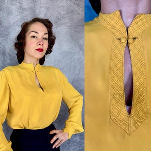 May include: Mustard yellow blouse with long sleeves and a mandarin collar. The collar and neckline feature intricate stitched detailing. The blouse has a keyhole opening at the neckline and is paired with a dark blue skirt. The sleeves have a slight puff at the shoulders and gather at the wrists.