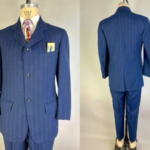 Puede incluir: Un traje azul de rayas diplom&aacute;ticas que incluye una chaqueta de tres botones con solapas de muesca, dos bolsillos con solapa y un bolsillo en el pecho con una tarjeta amarilla que muestra la ilustraci&oacute;n de un p&aacute;jaro azul. Visto de frente y de espaldas.