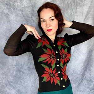 May include: Black cardigan sweater with a poinsettia floral design in red and green. The sweater has long sleeves and gold buttons. The background is a light gray.