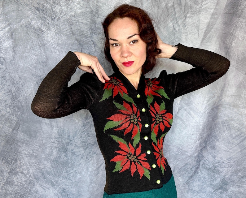 May include: Black cardigan sweater with a poinsettia floral design in red and green. The sweater has long sleeves and gold buttons. The background is a light gray.