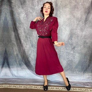 May include: A burgundy vintage dress with long sleeves and a midi-length skirt. The bodice features intricate embroidery, and a black belt cinches the waist. The dress is paired with black heels. The background is a neutral grey.