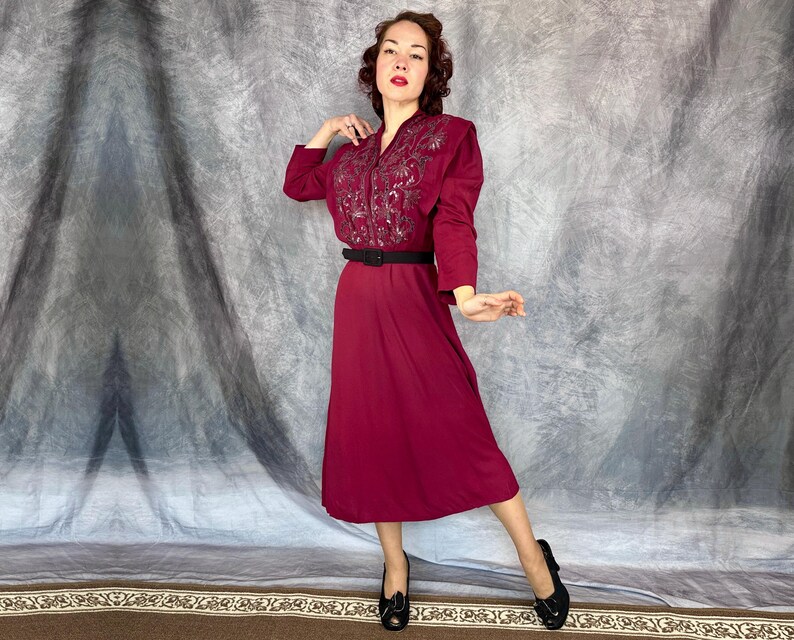 May include: A burgundy vintage dress with long sleeves and a midi-length skirt. The bodice features intricate embroidery, and a black belt cinches the waist. The dress is paired with black heels. The background is a neutral grey.