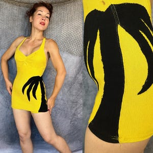 May include: A bright yellow sleeveless mini dress featuring a prominent black palm tree graphic on the side. The garment has a scoop neckline with subtle ruching at the center of the bust and a fitted silhouette that ends at the upper thigh.