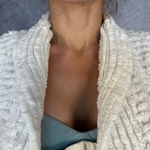 May include: A white textured jacket with a shawl collar featuring a shaggy, looped fabric construction. The garment has a thick, layered appearance with a soft, fuzzy surface texture throughout the front panels and collar, worn over a light blue top.