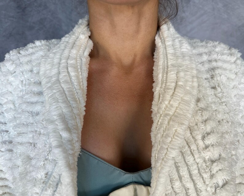 May include: A white textured jacket with a shawl collar featuring a shaggy, looped fabric construction. The garment has a thick, layered appearance with a soft, fuzzy surface texture throughout the front panels and collar, worn over a light blue top.