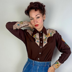 May include: A long-sleeved button-down shirt in dark brown with tan panels on the shoulders and cuffs. The tan sections feature colourful floral embroidery in yellow, blue, and green. The shirt has a pointed collar and white snap-style buttons.