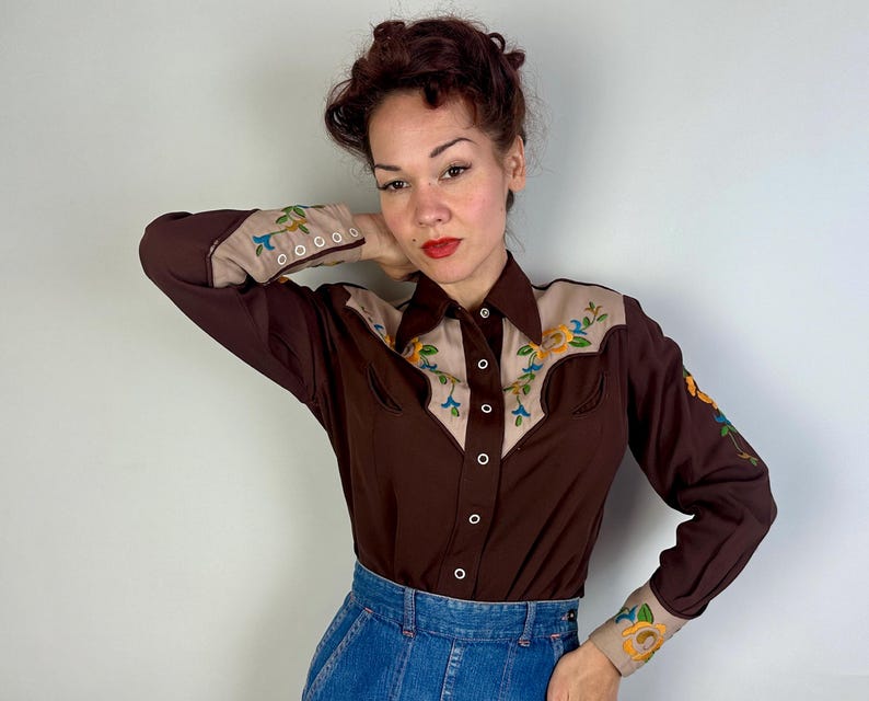 May include: A long-sleeved button-down shirt in dark brown with tan panels on the shoulders and cuffs. The tan sections feature colourful floral embroidery in yellow, blue, and green. The shirt has a pointed collar and white snap-style buttons.