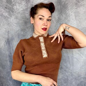 Pu&ograve; includere: Un top in maglia marrone con colletto e paramontura decorativi. Il top presenta un motivo a rombi sul davanti e maniche corte. Il colletto e la paramontura sono adornati da un motivo color crema. Il top &egrave; in stile vintage.