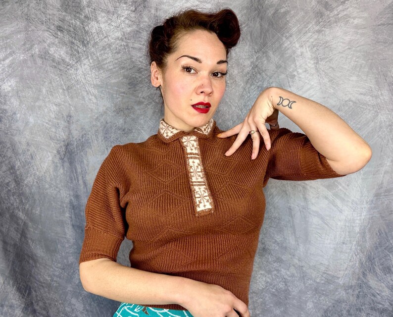 Pu&ograve; includere: Un top in maglia marrone con colletto e paramontura decorativi. Il top presenta un motivo a rombi sul davanti e maniche corte. Il colletto e la paramontura sono adornati da un motivo color crema. Il top &egrave; in stile vintage.