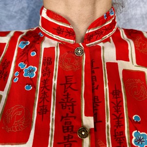 Pode incluir: Uma camisa de gola mandarim em vermelho e creme, com pain&eacute;is retangulares verticais contendo caligrafia asi&aacute;tica preta, motivos de grous dourados e pequenos detalhes florais azuis. A pe&ccedil;a inclui dois bot&otilde;es met&aacute;licos circulares na frente.