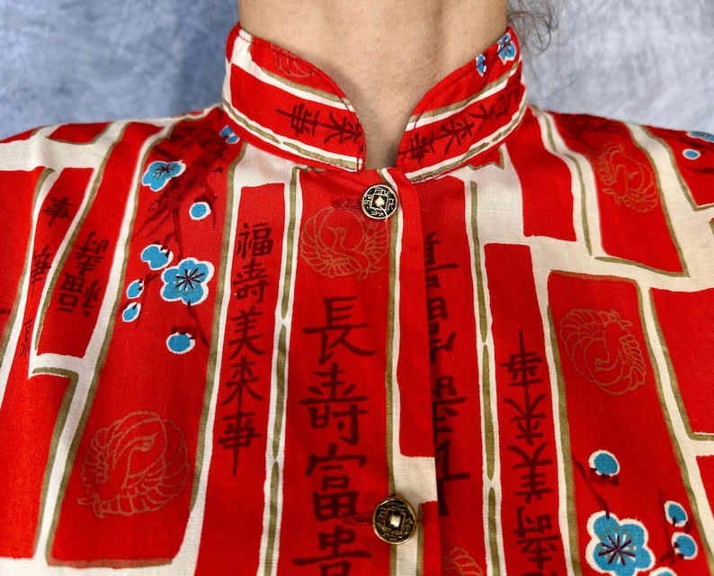 Pode incluir: Uma camisa de gola mandarim em vermelho e creme, com pain&eacute;is retangulares verticais contendo caligrafia asi&aacute;tica preta, motivos de grous dourados e pequenos detalhes florais azuis. A pe&ccedil;a inclui dois bot&otilde;es met&aacute;licos circulares na frente.