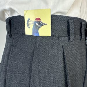 May include: High-waisted charcoal gray trousers with a textured diamond pattern, featuring belt loops and a side zipper closure. A small yellow rectangular card with an illustration of a peacock wearing a red bowler hat is tucked into the waistband.