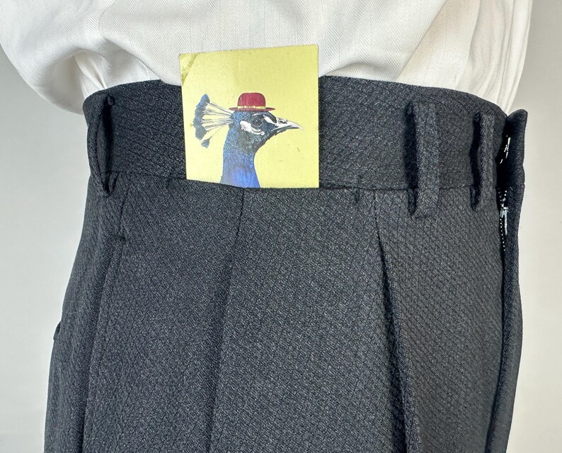 May include: High-waisted charcoal gray trousers with a textured diamond pattern, featuring belt loops and a side zipper closure. A small yellow rectangular card with an illustration of a peacock wearing a red bowler hat is tucked into the waistband.