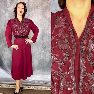 May include: A burgundy vintage dress with long sleeves and a V-neckline. The bodice features intricate sequin and embroidered detailing. A black belt cinches the waist, and the dress falls to a midi length. The dress is paired with black shoes.