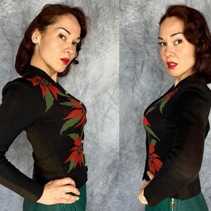 May include: A black knit cardigan with a poinsettia design in red and green. The long-sleeved jacket has a button-up front and is worn with a green skirt. The cardigan is a vintage style, perfect for the holiday season.