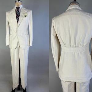 May include: A cream-coloured suit consisting of a two-button blazer and matching trousers. The blazer features a notched lapel, two front pockets, and a breast pocket containing a small card. A dark tie with a red and blue plaid pattern is worn.