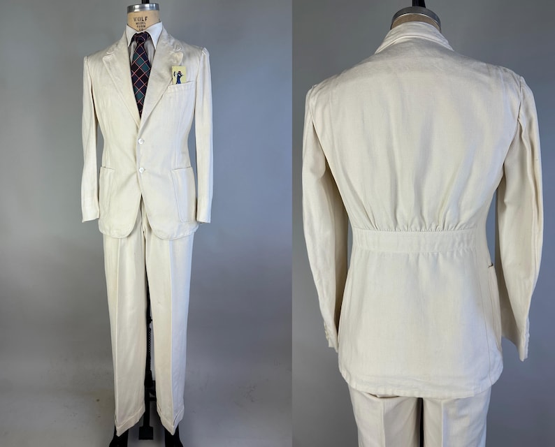 May include: A cream-coloured suit consisting of a two-button blazer and matching trousers. The blazer features a notched lapel, two front pockets, and a breast pocket containing a small card. A dark tie with a red and blue plaid pattern is worn.