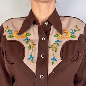May include: A brown long-sleeve shirt featuring beige panels on the chest and shoulders. The beige sections are decorated with embroidered floral patterns in yellow, green, and blue. The shirt has a pointed collar and white snap buttons.