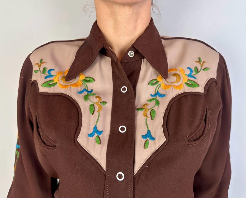 May include: A brown long-sleeve shirt featuring beige panels on the chest and shoulders. The beige sections are decorated with embroidered floral patterns in yellow, green, and blue. The shirt has a pointed collar and white snap buttons.