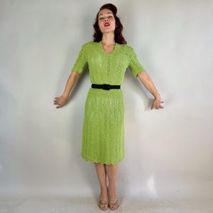 May include: A lime green knitted dress with short sleeves and a V-neck. The dress is cinched at the waist with a black belt. The hemline has a scalloped edge. The dress is knee-length.
