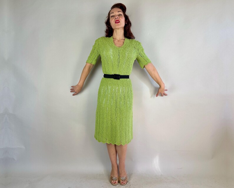 May include: A lime green knitted dress with short sleeves and a V-neck. The dress is cinched at the waist with a black belt. The hemline has a scalloped edge. The dress is knee-length.