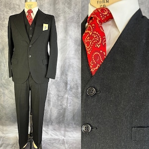 May include: A black three-piece suit consisting of a blazer, a vest, and trousers. The blazer features a two-button closure and a pocket square. A white collared shirt is worn underneath with a red necktie featuring a gold paisley pattern.