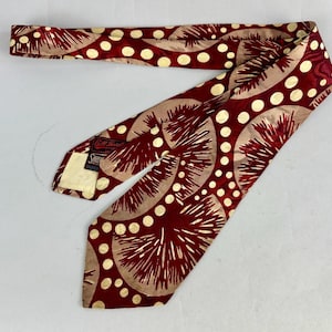 May include: A vintage necktie featuring a repeating pattern of beige and white circles and red spiky shapes. The tie has a label that reads "Bill Blass". The tie is made of a silky material.