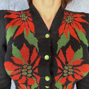 May include: Black cardigan sweater with a poinsettia floral design in red and green. The sweater has a V-neck, long sleeves, and green button closures. The floral pattern is repeated across the front of the sweater.