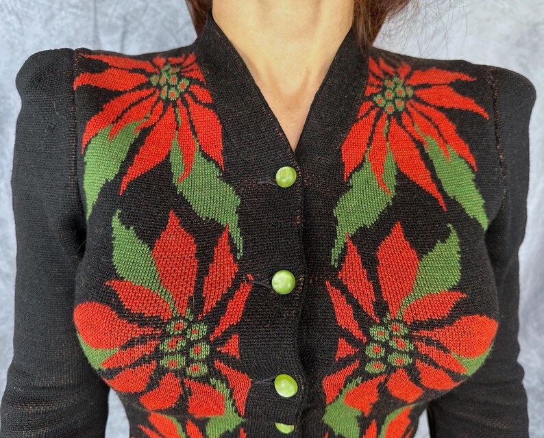 May include: Black cardigan sweater with a poinsettia floral design in red and green. The sweater has a V-neck, long sleeves, and green button closures. The floral pattern is repeated across the front of the sweater.