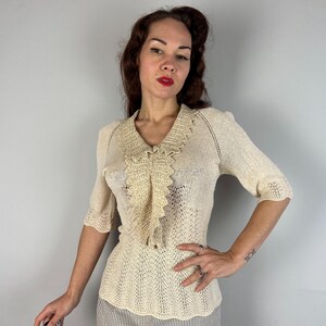 May include: A cream-colored crocheted top with a delicate lace collar and short sleeves. The top has a fitted silhouette and a delicate openwork pattern.