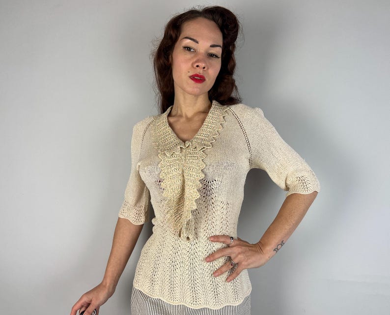 May include: A cream-colored crocheted top with a delicate lace collar and short sleeves. The top has a fitted silhouette and a delicate openwork pattern.