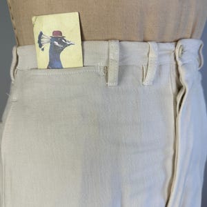 May include: Off-white canvas trousers featuring a high-waisted design with belt loops and a front button closure. A small rectangular card is tucked into the front pocket, displaying a blue peacock illustration wearing a small red bowler hat.
