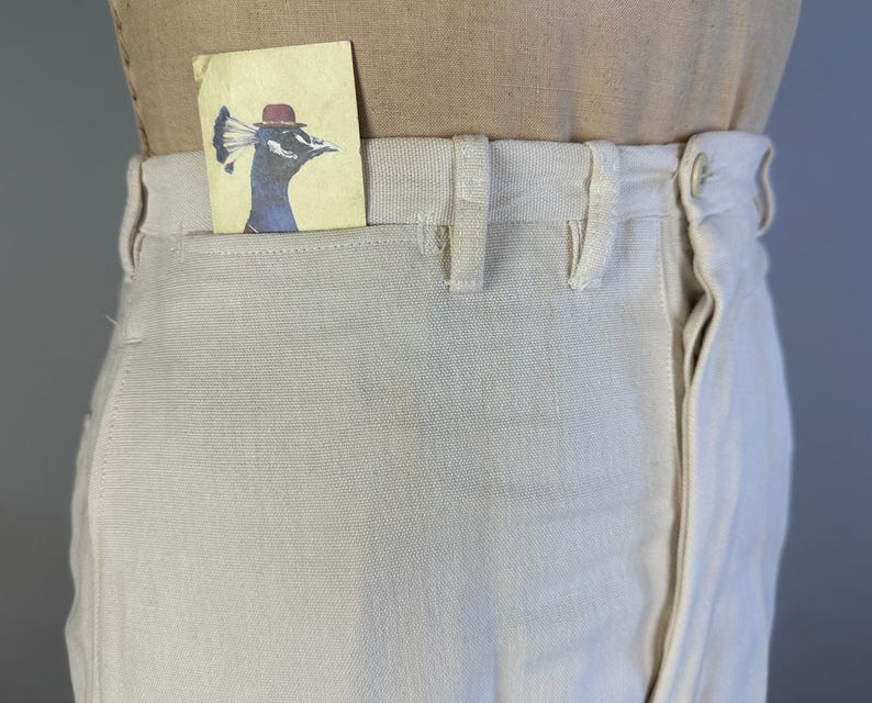 May include: Off-white canvas trousers featuring a high-waisted design with belt loops and a front button closure. A small rectangular card is tucked into the front pocket, displaying a blue peacock illustration wearing a small red bowler hat.