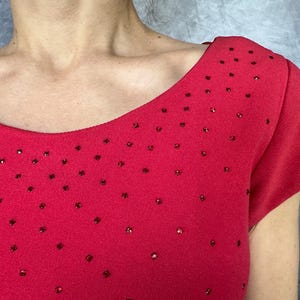May include: A vibrant red short-sleeved top featuring a wide, rounded neckline. The fabric is adorned with numerous small, scattered red rhinestones that create a subtle, sparkling effect across the front and shoulder areas of the garment.