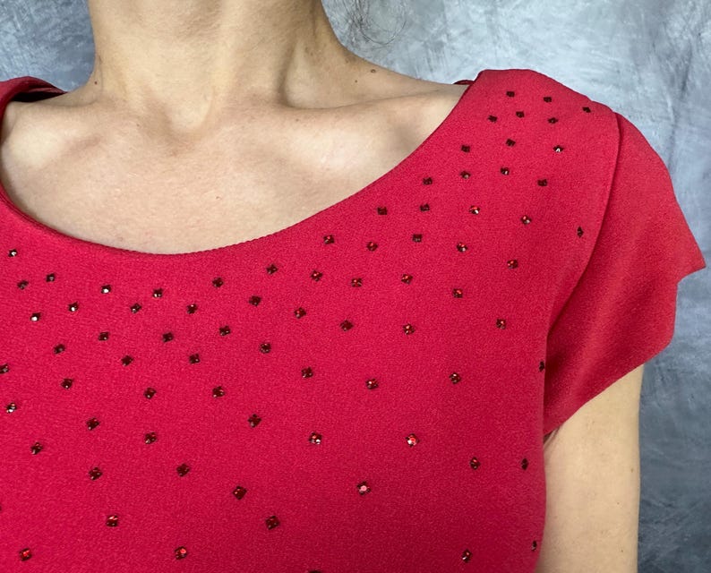 May include: A vibrant red short-sleeved top featuring a wide, rounded neckline. The fabric is adorned with numerous small, scattered red rhinestones that create a subtle, sparkling effect across the front and shoulder areas of the garment.