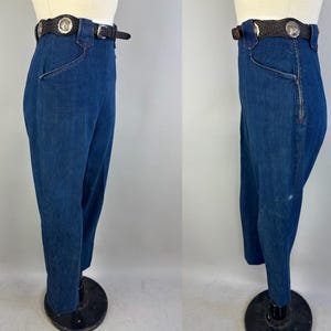 May include: High-waisted dark blue denim pants featuring a side zipper closure and a dark brown leather belt with silver-toned circular concho accents. The pants are displayed on a mannequin, showing both a front and side profile view of the garment.