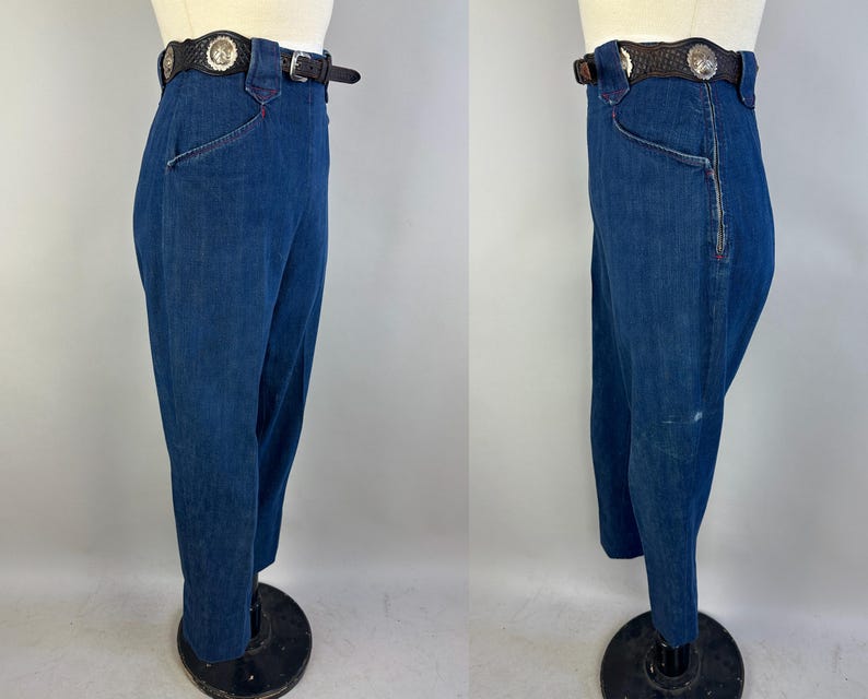 May include: High-waisted dark blue denim pants featuring a side zipper closure and a dark brown leather belt with silver-toned circular concho accents. The pants are displayed on a mannequin, showing both a front and side profile view of the garment.