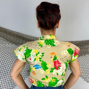 May include: Tan short-sleeved shirt with a tropical floral print featuring green leaves, yellow and white flowers, and cocktail illustrations. The shirt has a collared neckline and a button-down front. Text is visible on the shirt.