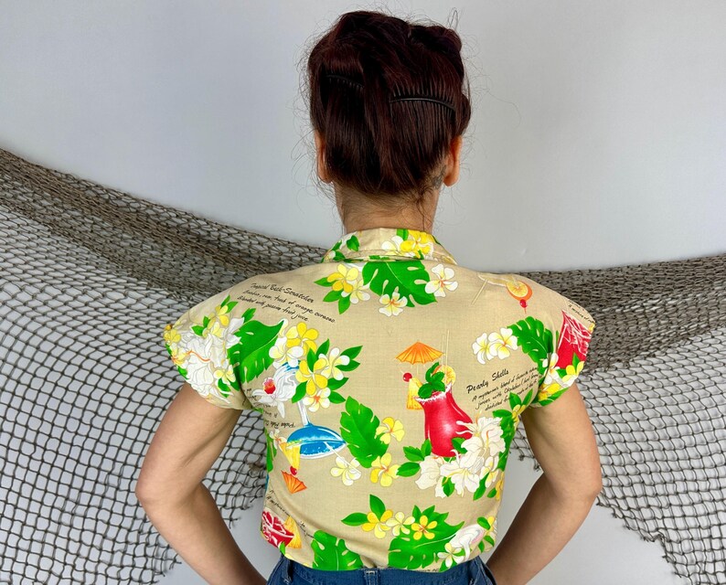 May include: Tan short-sleeved shirt with a tropical floral print featuring green leaves, yellow and white flowers, and cocktail illustrations. The shirt has a collared neckline and a button-down front. Text is visible on the shirt.