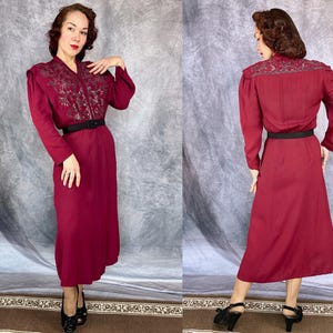 May include: A burgundy dress with long sleeves and a black belt. The dress features intricate embroidery on the shoulders and bodice. The dress is knee-length and has a classic, vintage style.