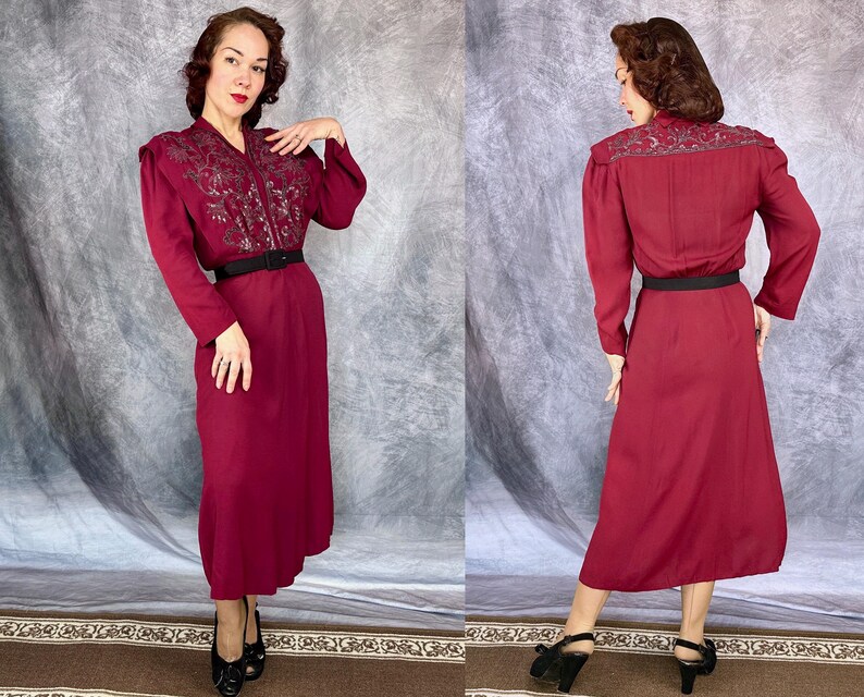 May include: A burgundy dress with long sleeves and a black belt. The dress features intricate embroidery on the shoulders and bodice. The dress is knee-length and has a classic, vintage style.