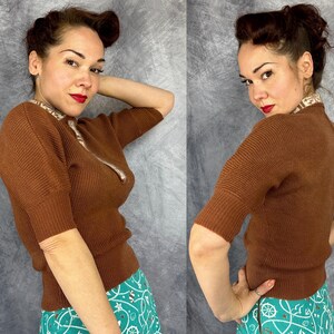 Pu&ograve; includere: Un maglione marrone a maniche corte con una trama a coste e una cerniera. Il maglione ha un colletto fantasia ed &egrave; abbinato a una gonna turchese con un disegno bianco e verde acqua. La persona indossa il rossetto rosso.